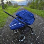 Teel seisab thule lapsevanker, millel on sinine vihmakile. Taga on rohelised puud ja rohi.