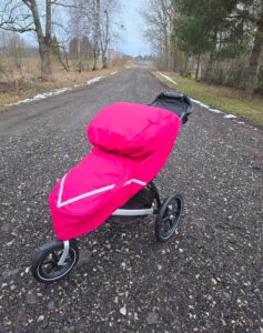 Teel seisab lapsevanker, millel on roosa lukuga vihmakile. Tee ääres on natuke lund. Puud on raagus.